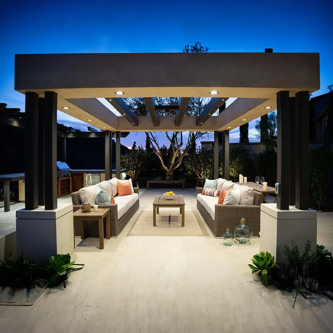 Outdoor patio with wicker sofas under a pergola, illuminated at dusk. The setup includes beige cushioned seating, a wooden coffee table, and side tables, creating a cozy ambiance with landscape lighting