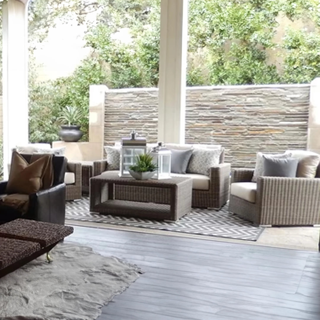 Stylish outdoor patio area featuring wicker furniture with beige cushions, a coffee table with lanterns and potted plants, and a textured stone wall with lush greenery in the background