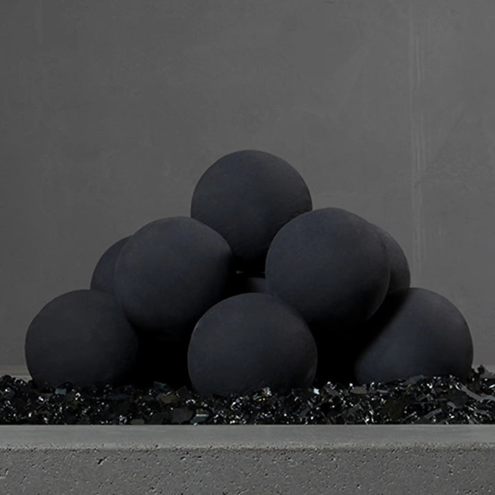 Close-up view of Starfire Designs 4-inch ceramic fire balls set of 12 in dark gray, stacked in a pyramid on a bed of black fire glass in an unlit fire feature. Their smooth texture and modern style enhance any contemporary fire pit setup