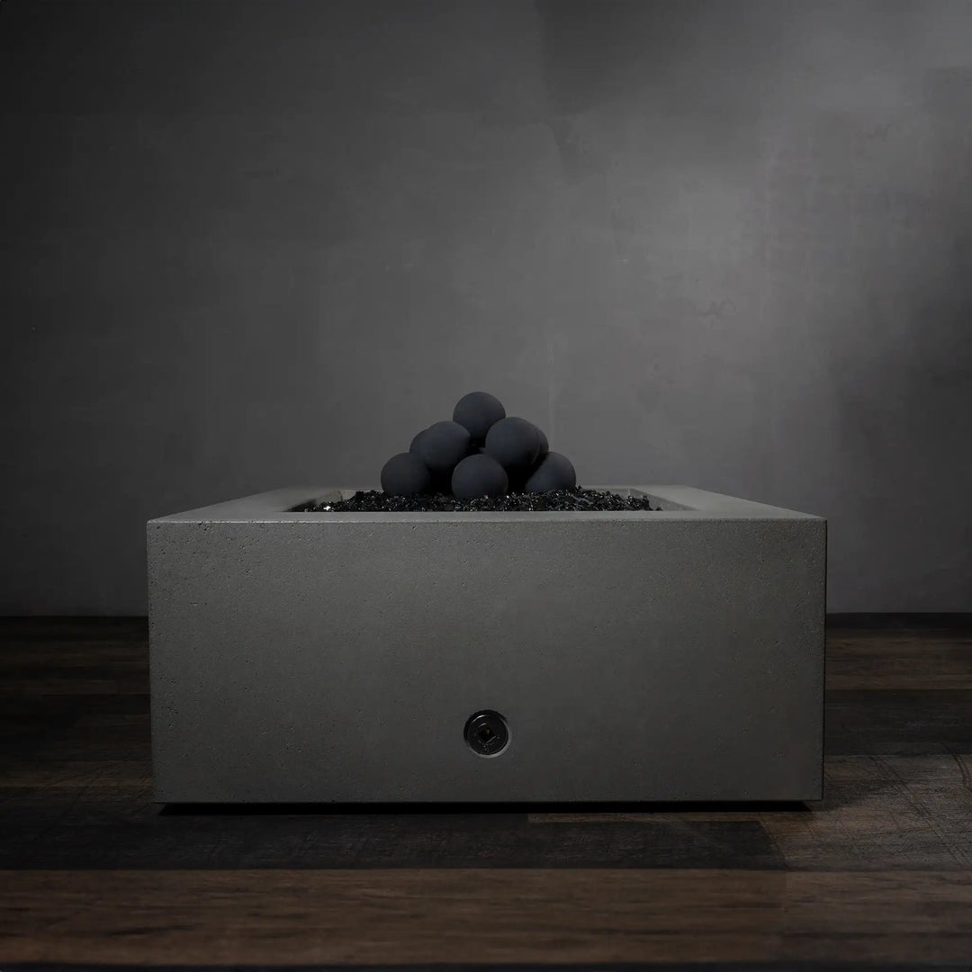 Starfire Designs 4-inch ceramic fire balls set of 12 in dark gray resting on top of a bed of black fire glass in an unlit square gas fire pit, providing a contemporary, sleek appearance even without flames. Ideal for modern fire pit designs