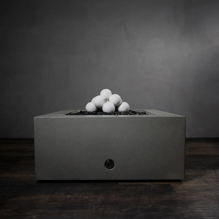 Set of 12 white 4-inch ceramic fire balls from Starfire Designs displayed in a pyramid on top of black fire glass in an unlit square concrete fire pit. The minimalist design of the fire balls enhances the sleek aesthetic of the fire pit, making it a stylish addition to outdoor areas. Durable and heat-resistant, these ceramic fireballs are perfect for gas fire pits in both residential and commercial applications. White fire pit decoration, outdoor gas fire pit accessory, modern fire décor