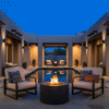 In a rectangular courtyard at night, a dark graphite patio fire pit sits on flagstone near the edge of a long reflecting pool, its cylindrical concrete fire pit body centered between two cushioned armchairs with patterned pillows, while warm wall sconces and a second smaller flame echo the same fire feature at the far end of the space, giving the symmetrical layout the appearance of a family gathering fire pit area framed by stucco walls and sparse desert plants.
