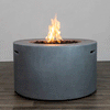 A cylindrical gray round fire pit sits on a wood-look floor against a plain backdrop, its thick concrete fire pit walls enclosing a recessed bed of lava rocks where a tall, steady flame burns from a hidden burner, emphasizing the clean, sculptural lines of this modern fire pit; the design suggests a low-maintenance gas fire pit ideal for contemporary patios, rooftop lounges, or courtyard seating areas that call for understated warmth and style.