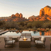 On a raised wood deck beside a still pool and desert landscaping, a light sand patio fire pit shaped as a square fire pit sits between a cushioned loveseat and two matching armchairs, its center filled with dark lava rocks and a small orange flame, while agave plants, shrubs, and red rock formations in the distance frame the outdoor fire pit against a pale evening sky.
