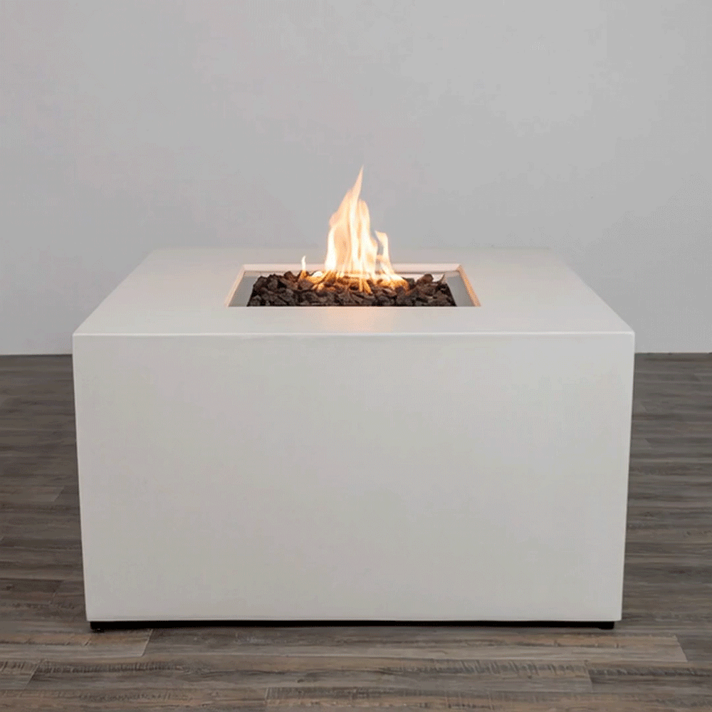 In a plain studio-style setting, a solid white concrete fire pit with straight sides stands on a dark wood floor, the piece shaped as a low cube with a centered square fire pit opening cut into the top surface, where jagged dark lava rocks fill the recess and a steady orange flame rises, the clean edges, smooth finish, and lack of additional objects emphasizing a simple modern fire pit presented alone against a light wall.