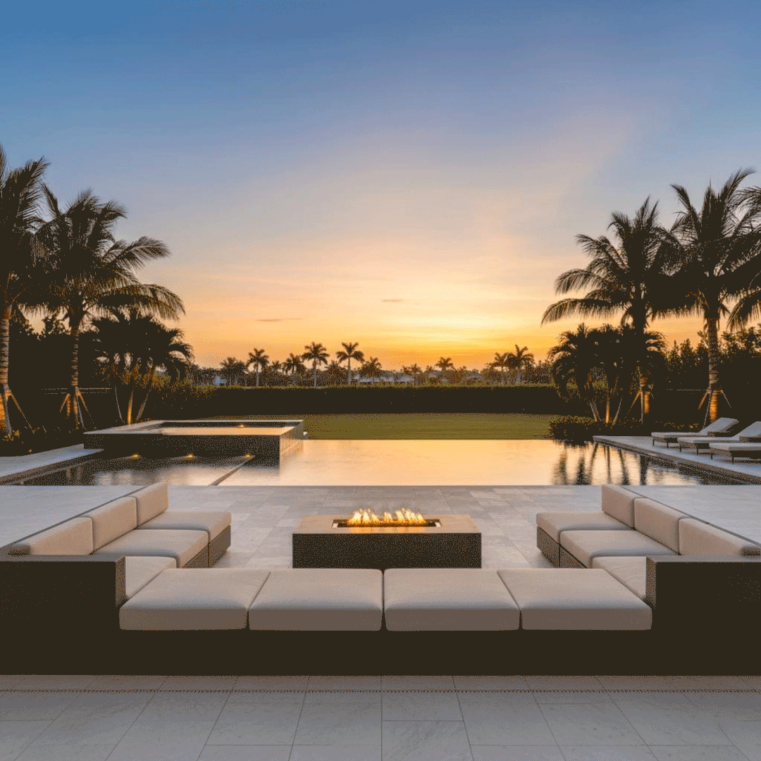 A pair of low cushioned sectionals face each other across a gray rectangular fire pit on a wide tile terrace, the central fire feature showing a row of flames above dark stones at the edge of an infinity pool, with palm trees, lawn, and the warm glow of sunset forming a backdrop that makes the poolside arrangement resemble a resort-like backyard oasis prepared for relaxed outdoor entertaining.