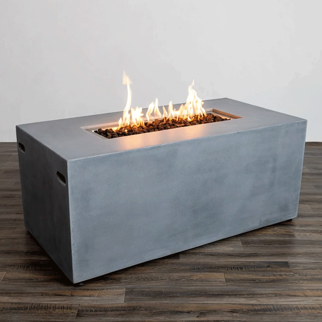 In a simple studio setting, a natural gray rectangular fire pit with straight sides and subtle handholds rests on a dark wood plank floor against a pale wall, the smooth surface identifying it as a concrete fire pit while a long recessed opening runs along the top center, holding uneven black lava rocks and a continuous row of bright flames, the clean geometry and lack of other objects emphasizing a minimal modern fire pit shape.