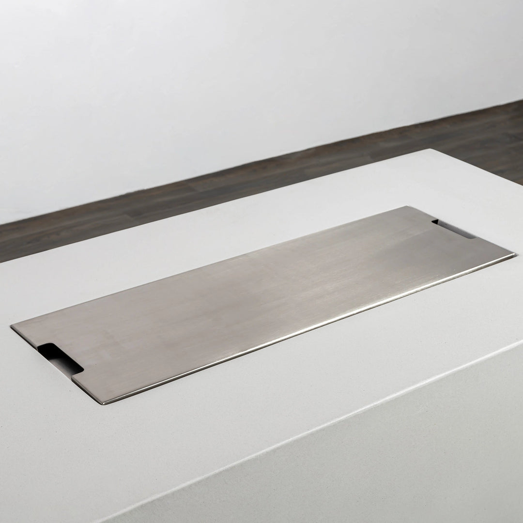 A close-up of the Starfire Designs Beton 55" White Rectangle Hidden Propane Tank Gas Fire Pit with the stainless steel burner cover in place. The cover sits flush on the rectangular concrete frame, offering a clean and minimalist aesthetic. This protective cover is perfect for shielding the burner when not in use.