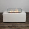 This image highlights the Starfire Designs Beton 55" White Rectangle Hidden Propane Tank Gas Fire Pit with the glass wind guard installed and the flames ignited. The bright, vibrant flames dance over the lava rock filler, creating a warm ambiance. The tempered glass guard enhances safety while adding a stylish accent.