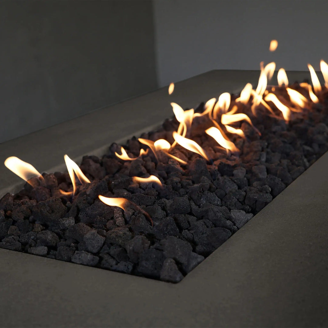A close-up view of the Starfire Designs Concrete Rustic 60" x 32" Rectangle Gravity Gas Fire Pit in brown, focusing on the glowing flames dancing above black lava rocks. The natural texture of the concrete contrasts beautifully with the fire and rocks, creating a captivating scene. This fire pit is ideal for bringing warmth and a rustic touch to modern outdoor environments.