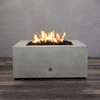 A close-up studio view presents a square fire pit cast in smooth grey concrete, resting on a dark wood floor against a matching wall. Bright yellow flames burn continuously from a bed of black lava rocks, showcasing the clean combustion and surface texture of the concrete fire pit. A chrome key valve is visible on the side panel, indicating the manual control mechanism of this natural gas fire pit unit.