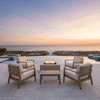 A white patio fire pit anchors a seating area with four cushioned armchairs, positioned on a tiled deck overlooking beach dunes and the ocean. The golden sunset light complements the warm amber flames of the propane fire pit, creating a cozy backyard fire pit atmosphere. The clean burn demonstrates a smokeless fire pit experience suitable for breezy coastal environments.