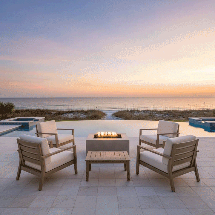 A white patio fire pit anchors a seating area with four cushioned armchairs, positioned on a tiled deck overlooking beach dunes and the ocean. The golden sunset light complements the warm amber flames of the propane fire pit, creating a cozy backyard fire pit atmosphere. The clean burn demonstrates a smokeless fire pit experience suitable for breezy coastal environments.
