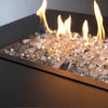 A stunning close-up of Starfire Glass 1/2" Broken Fire Glass (10lbs) in Titanium Reflective, glowing with a warm fire over a long rectangular gas fire pit. The premium crushed glass pieces reflect the flames, adding a modern, luxurious touch to any outdoor fire pit, fire bowl, or fire table setup.