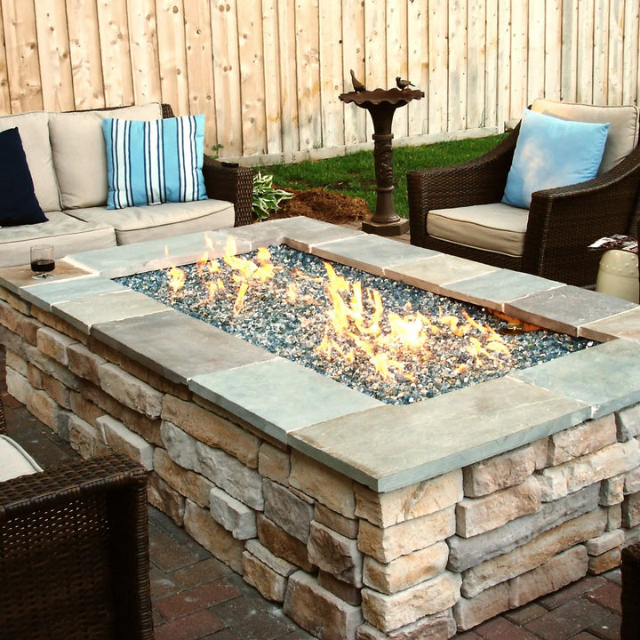 A rectangular stone fire pit table filled with Starfire Glass 1/2" Broken Pre-Mixed Fire Glass (10lbs), burning with vibrant flames. The reflective fire glass enhances the outdoor fire pit ambiance, surrounded by comfortable patio seating. Ideal for fire pit tables, backyard fire pits, and outdoor fire bowls.