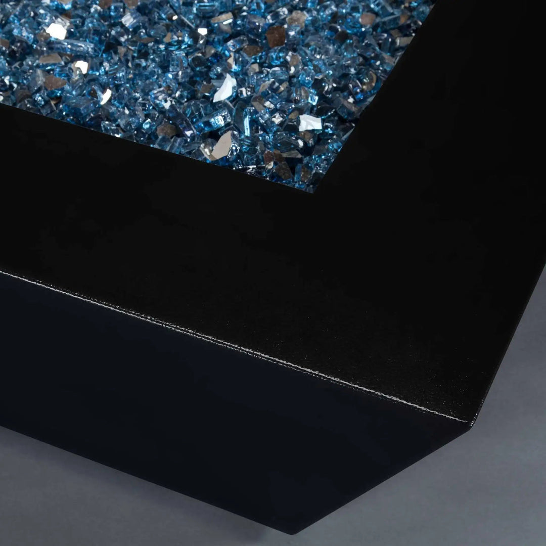 A close-up of the corner of the Steel Mill 48-inch by 32-inch Rectangle Gas Fire Pit - Black Hammertone, featuring reflective blue fire glass, enhancing the modern design
