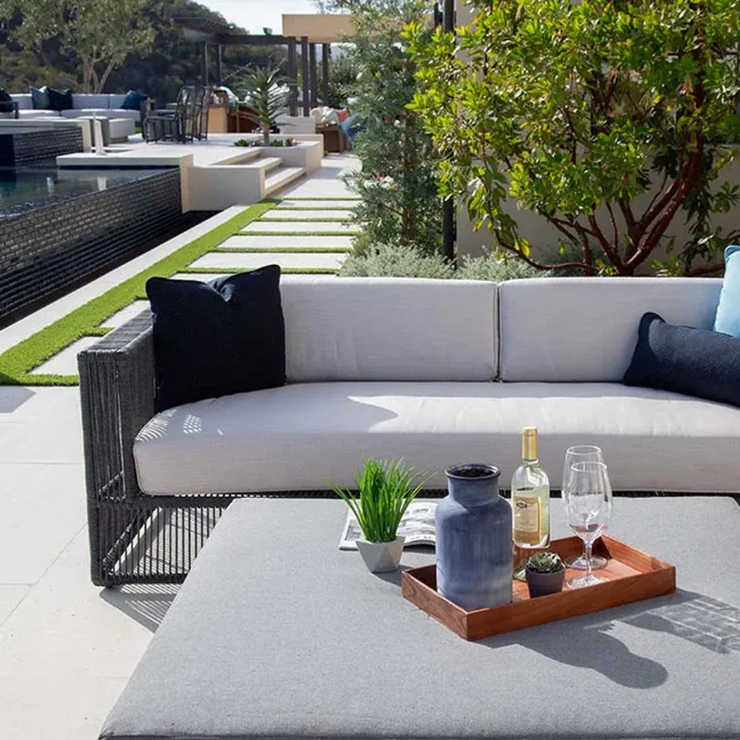 Stylish outdoor patio setup featuring a light-colored sofa with dark accent pillows, accompanied by a coffee table with decorative items, overlooking a pool and garden pathway