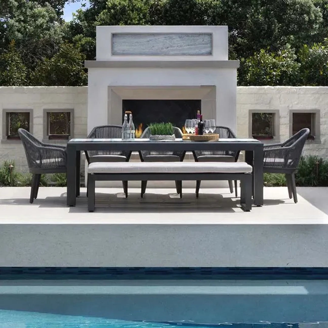 Modern outdoor dining set with a rectangular table, cushioned bench, and woven armchairs placed in front of a contemporary fireplace, with a pool in the foreground and greenery in the background