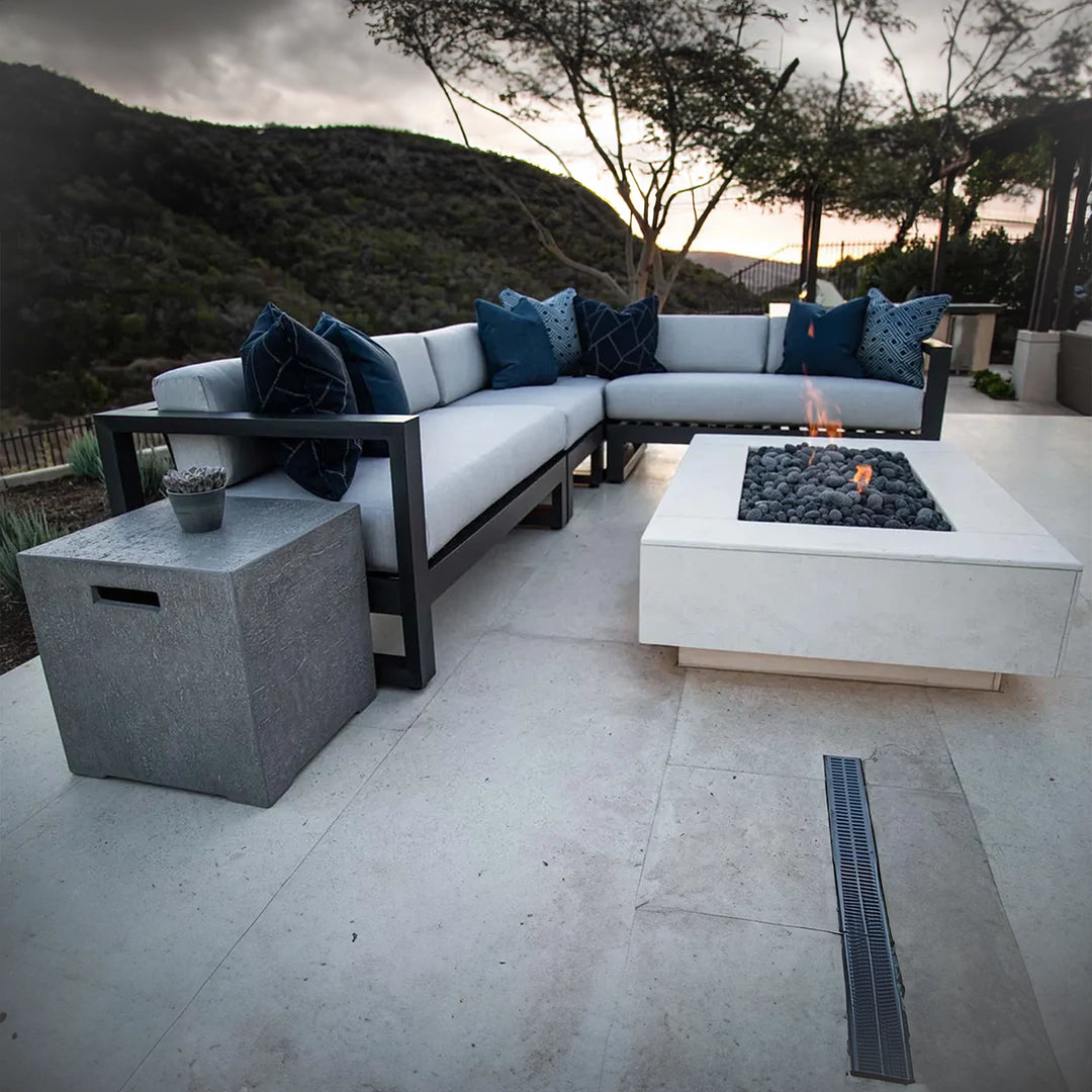 Modern outdoor sectional sofa with light cushions and dark blue pillows, surrounding a square fire pit with lava rocks on a patio overlooking natural hills.