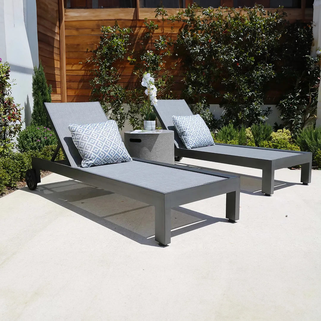 Pair of modern outdoor chaise lounges with light gray sling fabric and patterned pillows, arranged around a minimalist side table with an orchid on a sunny patio.