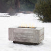The Outdoor Plus 108" Catalina Wood Grain Concrete Gas Fire Pit in ivory is featured in a snow-covered outdoor scene, illustrating its use in colder climates. The ivory wood grain texture stands out beautifully against the snow, creating a warm focal point in any landscape.