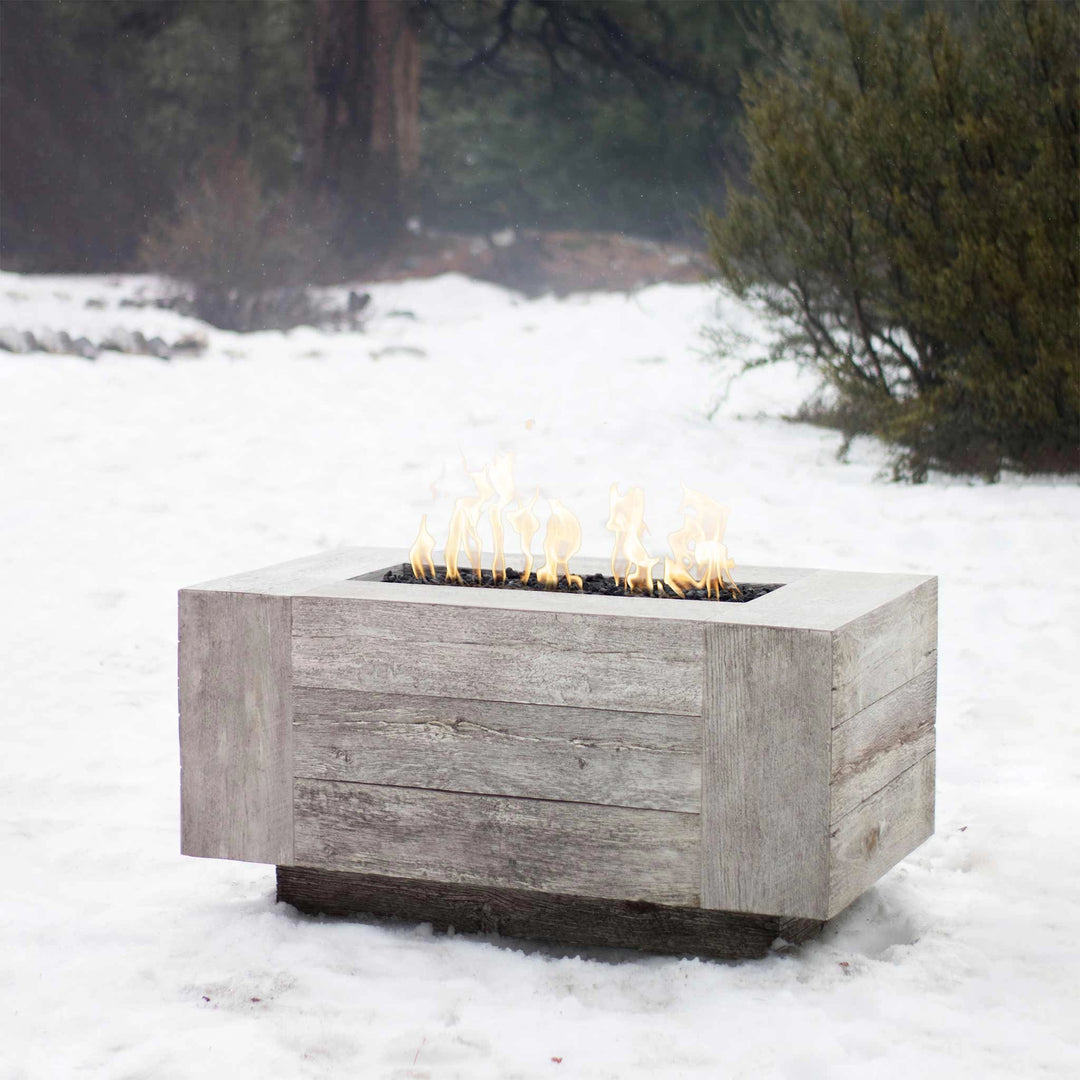 The Outdoor Plus 108" Catalina Wood Grain Concrete Gas Fire Pit in ivory is featured in a snow-covered outdoor scene, illustrating its use in colder climates. The ivory wood grain texture stands out beautifully against the snow, creating a warm focal point in any landscape.