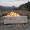 The Outdoor Plus 108" Coronado Wood Grain Concrete Gas Fire Pit in ivory set in a backyard patio, surrounded by wooden Adirondack chairs. The fire pit's wood grain texture complements the natural wood furniture, creating a cozy and inviting outdoor gathering space. The gas flames add warmth and ambiance, perfect for evening gatherings.