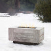 The Outdoor Plus 120-inch Catalina Wood Grain Concrete Gas Fire Pit in Ivory in a winter snow setting. Its rustic design with wood grain texture contrasts beautifully against the snowy backdrop, making it a stunning feature for year-round outdoor use, even in colder climates. The flames stand out against the white snow, creating a warm and inviting atmosphere