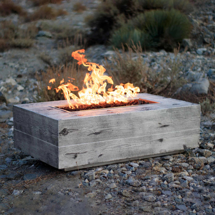 The Outdoor Plus 120-inch Coronado wood grain concrete gas fire pit in ivory, placed in an outdoor natural setting. The fire pit blends rustic charm with modern functionality, featuring a realistic wood grain texture in concrete and a large flame. Perfect for enhancing luxury outdoor environments with warmth and style