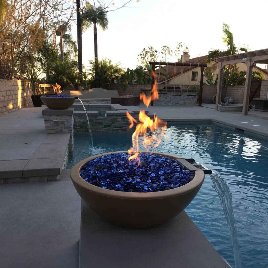 A serene outdoor poolside scene with two The Outdoor Plus 27" Round Concrete Sedona Fire and Water Bowls featuring vibrant blue fire glass and smooth waterfall accents. The fire and water bowl creates a captivating ambiance with its striking flames and calming water flow, perfectly suited for luxurious backyard pools or modern outdoor spaces. This design brings a sophisticated touch, blending contemporary style with functional beauty.