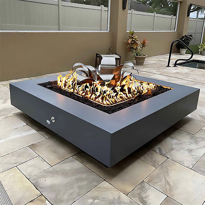 This lifestyle image showcases The Outdoor Plus 36-inch Cabo Square Concrete Gas Fire Pit in gray finish, installed on a modern patio. The sleek design complements the outdoor seating area, surrounded by pavers, while the fire pit is ablaze, providing warmth and ambiance. A stainless steel control valve is visible on the side, and black lava rocks fill the fire pit. Ideal for contemporary backyard and outdoor setups, this fire pit creates a luxurious and functional focal point.