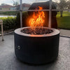 The Outdoor Plus 42-inch Chat Height Florence Concrete Gas Fire Pit in use during an evening setting. The fire pit emits a warm glow with flames dancing over dark fire glass. The sleek concrete design in a darker shade complements the modern outdoor space, making it perfect for relaxing by the fire during cool nights. This gas fire pit is an excellent addition for those seeking both a stylish and functional centerpiece for their outdoor area