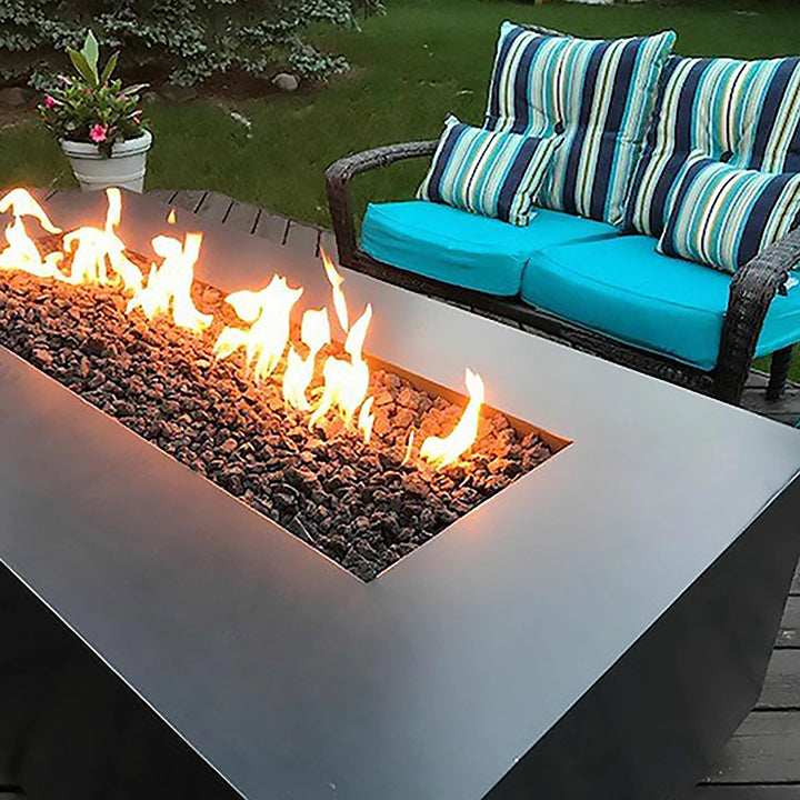 The Outdoor Plus 48" Coronado Steel Gas Fire Pit situated in a garden setting with vibrant greenery and comfortable outdoor seating. This sleek fire pit serves as a sophisticated focal point for the space, offering a blend of function and style. The radiant flames create a cozy and inviting atmosphere, making it an ideal fire feature for enjoying evenings outdoors.