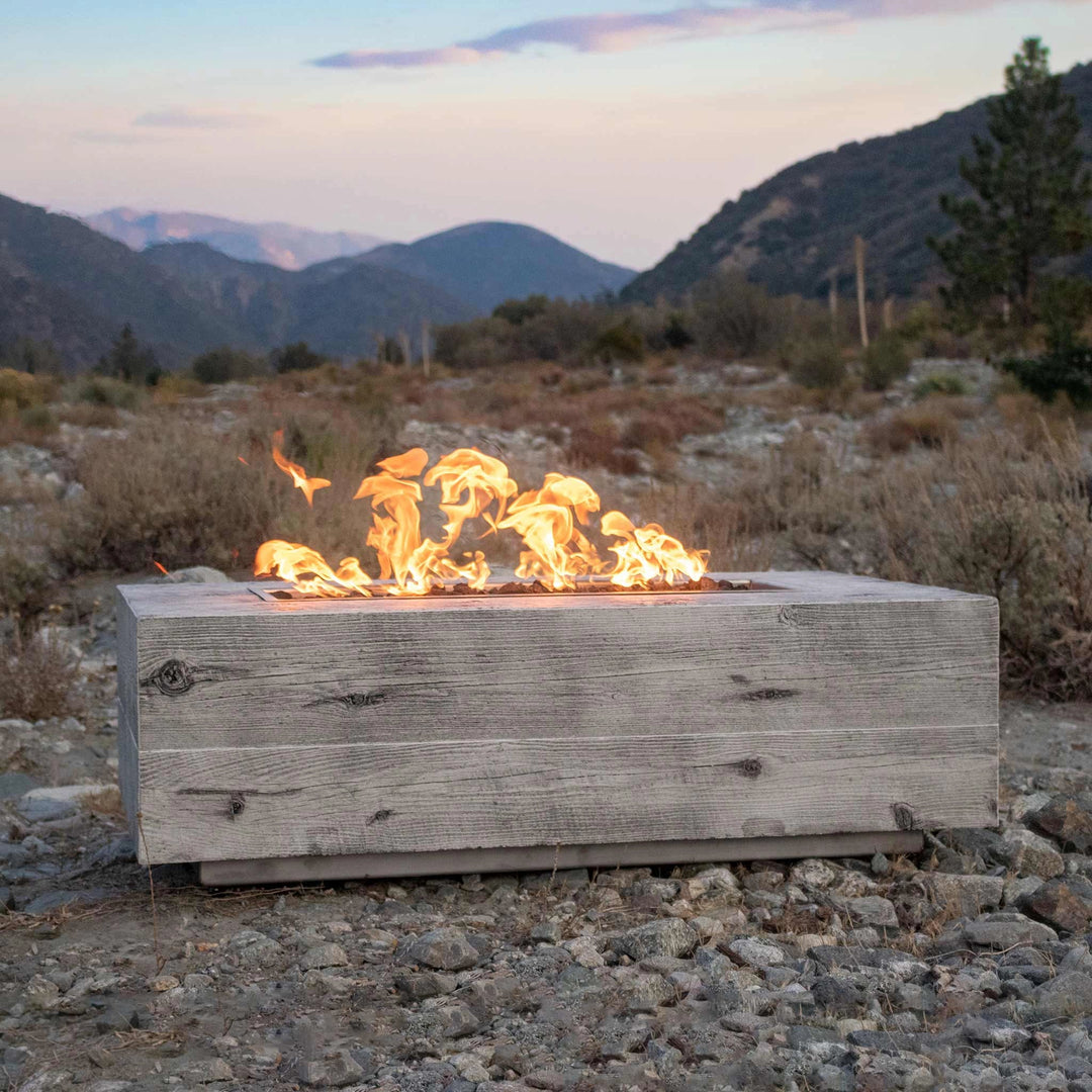 The Outdoor Plus 48" Coronado Wood Grain Concrete Gas Fire Pit placed in a rustic mountainous landscape, with vibrant flames contrasting against the natural backdrop of rocky terrain and distant mountains. The fire pit's wood grain texture in a light gray finish complements the rugged outdoor setting, making it an ideal choice for nature-inspired outdoor areas. A perfect addition for creating a warm gathering space. Outdoor gas fire pit designed for rustic and natural-themed settings