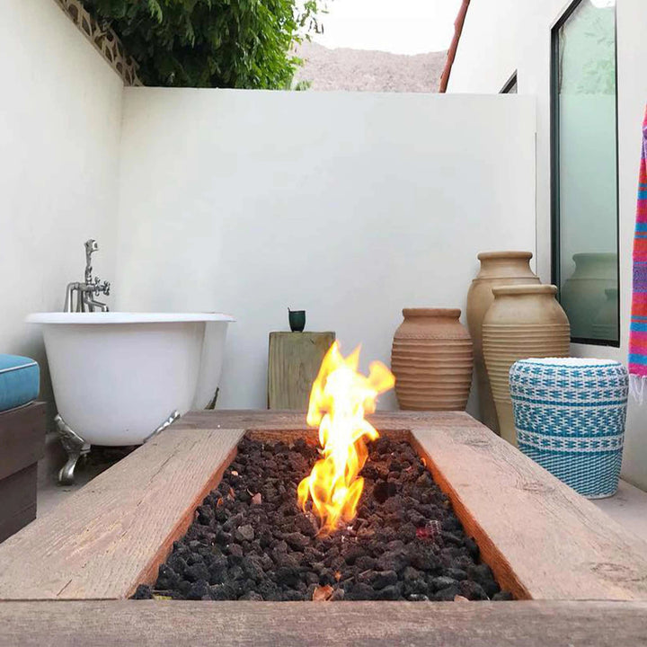 The Outdoor Plus 48" Coronado Wood Grain Concrete Gas Fire Pit set in a modern backyard oasis, blending rustic appeal with contemporary surroundings. The fire pit is displayed in a cozy outdoor patio area featuring a white bathtub, colorful accents, and decorative pottery, showcasing its versatility in design aesthetics. Perfect for creating a warm ambiance, this fire pit complements various backyard styles. Ideal for those seeking a stylish gas fire pit with a realistic wood grain texture