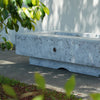 The Outdoor Plus 60-inch Del Mar Concrete Gas Fire Pit displayed outdoors with a rustic textured finish. This gas fire pit combines durability with a stylish design, ideal for enhancing any patio or backyard space with its rugged and elegant appearance. A modern solution for outdoor warmth and ambiance.