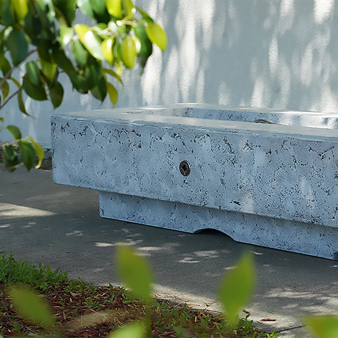 The Outdoor Plus 60-inch Del Mar Concrete Gas Fire Pit displayed outdoors with a rustic textured finish. This gas fire pit combines durability with a stylish design, ideal for enhancing any patio or backyard space with its rugged and elegant appearance. A modern solution for outdoor warmth and ambiance.