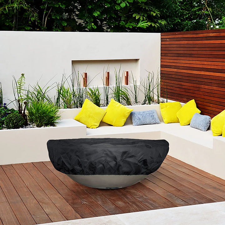 Lifestyle image showcasing "The Outdoor Plus Round Firepit Cover" in a modern patio setting. The round fire pit cover is placed on a large fire bowl, surrounded by a stylish patio arrangement with bright, decorative pillows. The cover is designed to blend seamlessly into outdoor aesthetics while providing complete protection against weather elements, including rain, wind, and UV rays. Perfect for maintaining the beauty of outdoor fire pits, fire bowls, and fire tables