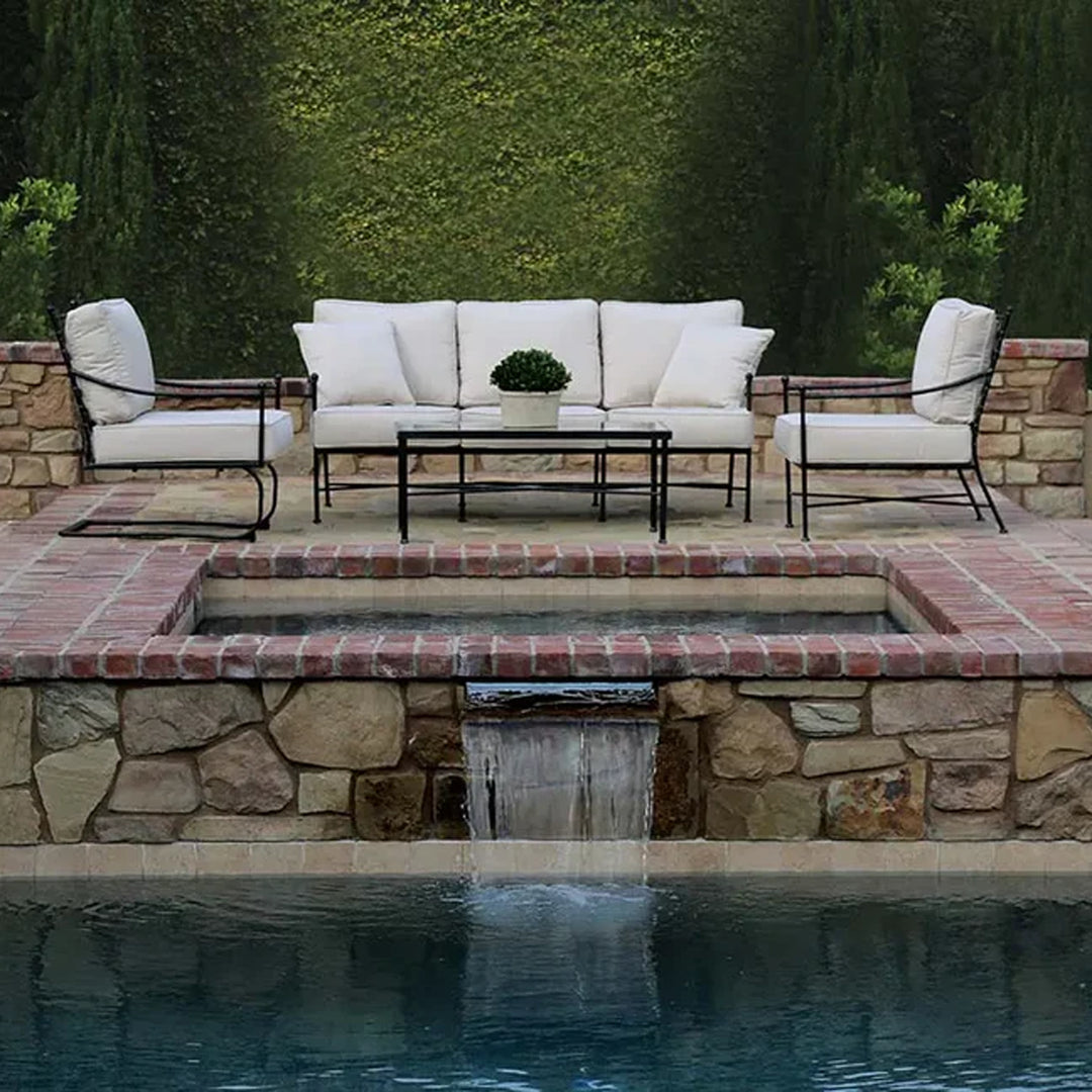 Outdoor patio featuring a metal frame sofa and chairs with white cushions, arranged on a brick and stone platform with a water feature and waterfall into a pool