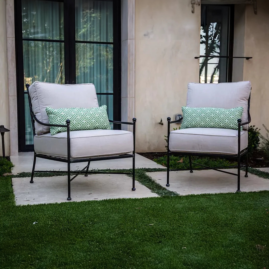 Two metal frame lounge chairs with white cushions and green decorative pillows on a stone patio, set beside a grassy area and modern house with large windows